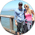 Man wearing sunglasses and woman with blonde hair posing by a wooden railing near water.