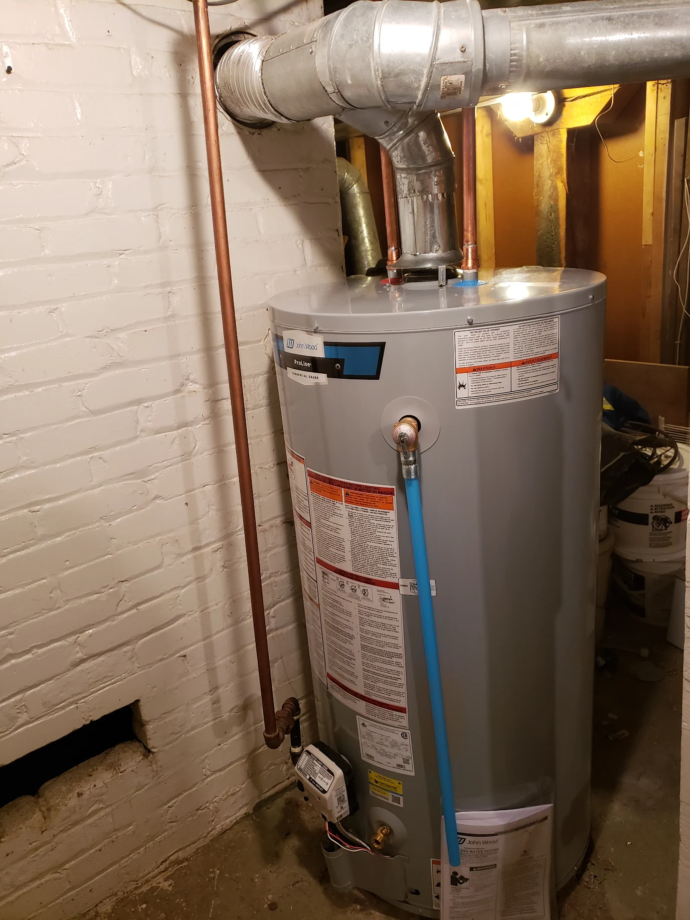Gray John Wood ProLine water heater with pipes and ductwork in a basement corner.