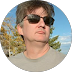 Man wearing sunglasses and a beige shirt against a blue sky with some autumn trees.