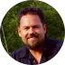 Smiling man with dark hair and beard wearing a dark-colored shirt, outdoors with greenery.
