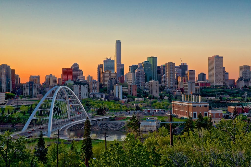 City skyline at sunset with modern skyscrapers, a white arched bridge, and river in the foreground.