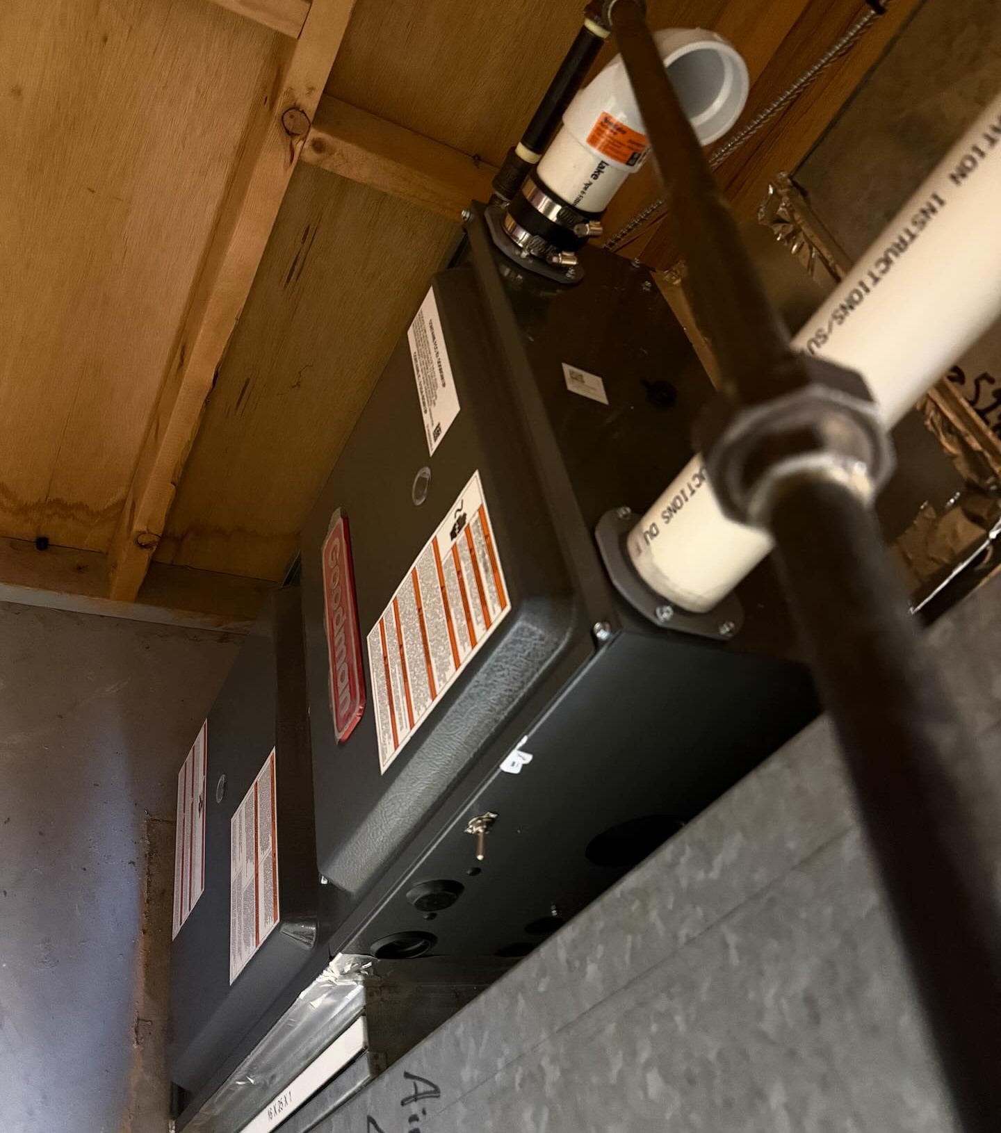 Two black Goodman HVAC units installed under a wooden ceiling with visible pipes connected.