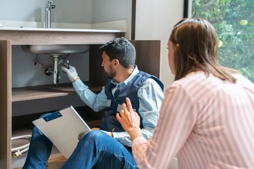 Plumber checking sink pipes under a kitchen cabinet while a woman watches and talks.