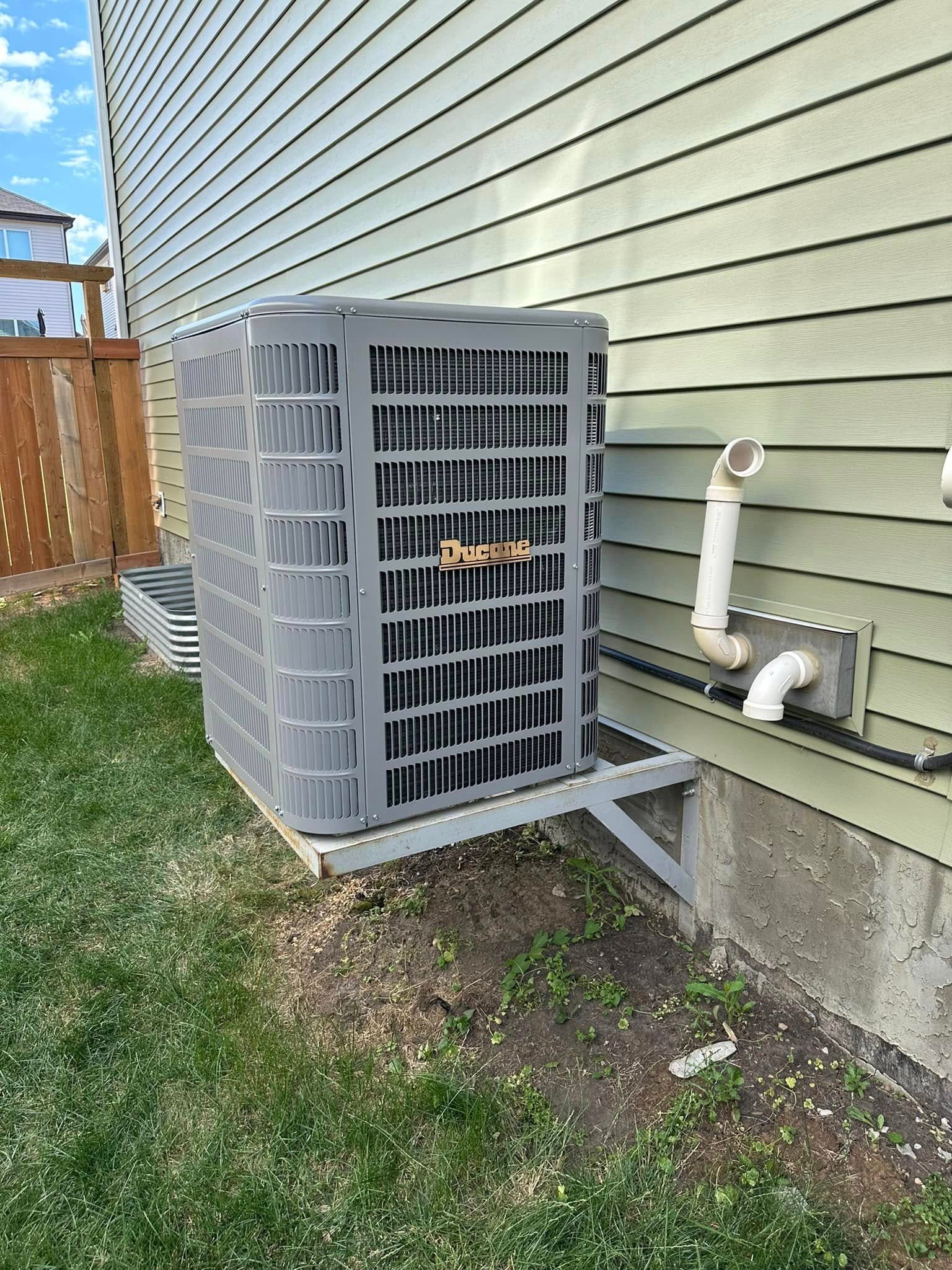 Ducane outdoor air conditioning unit mounted on metal bracket beside house with grass and wooden fence.