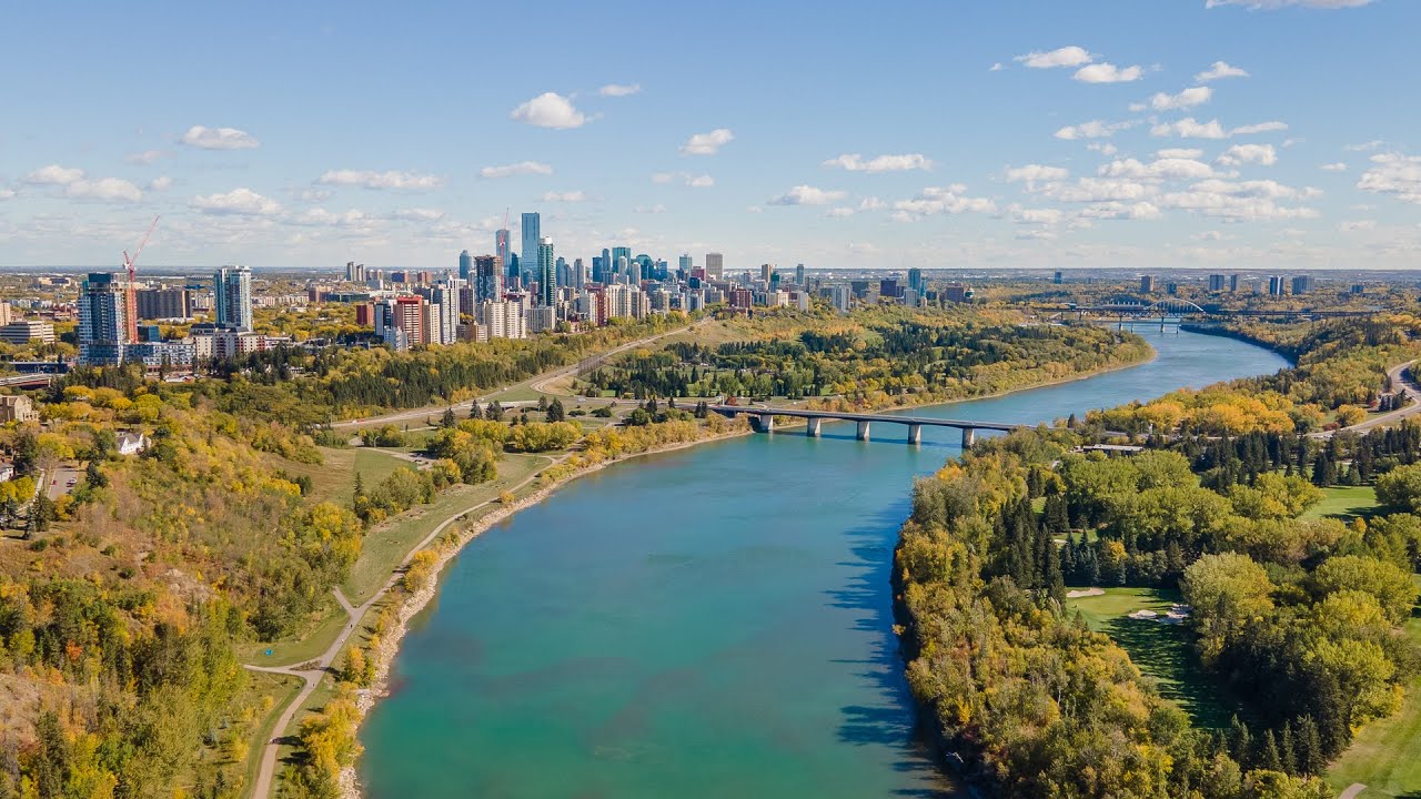 Wide river winding through green forest with city skyline and bridges under a blue sky with clouds.