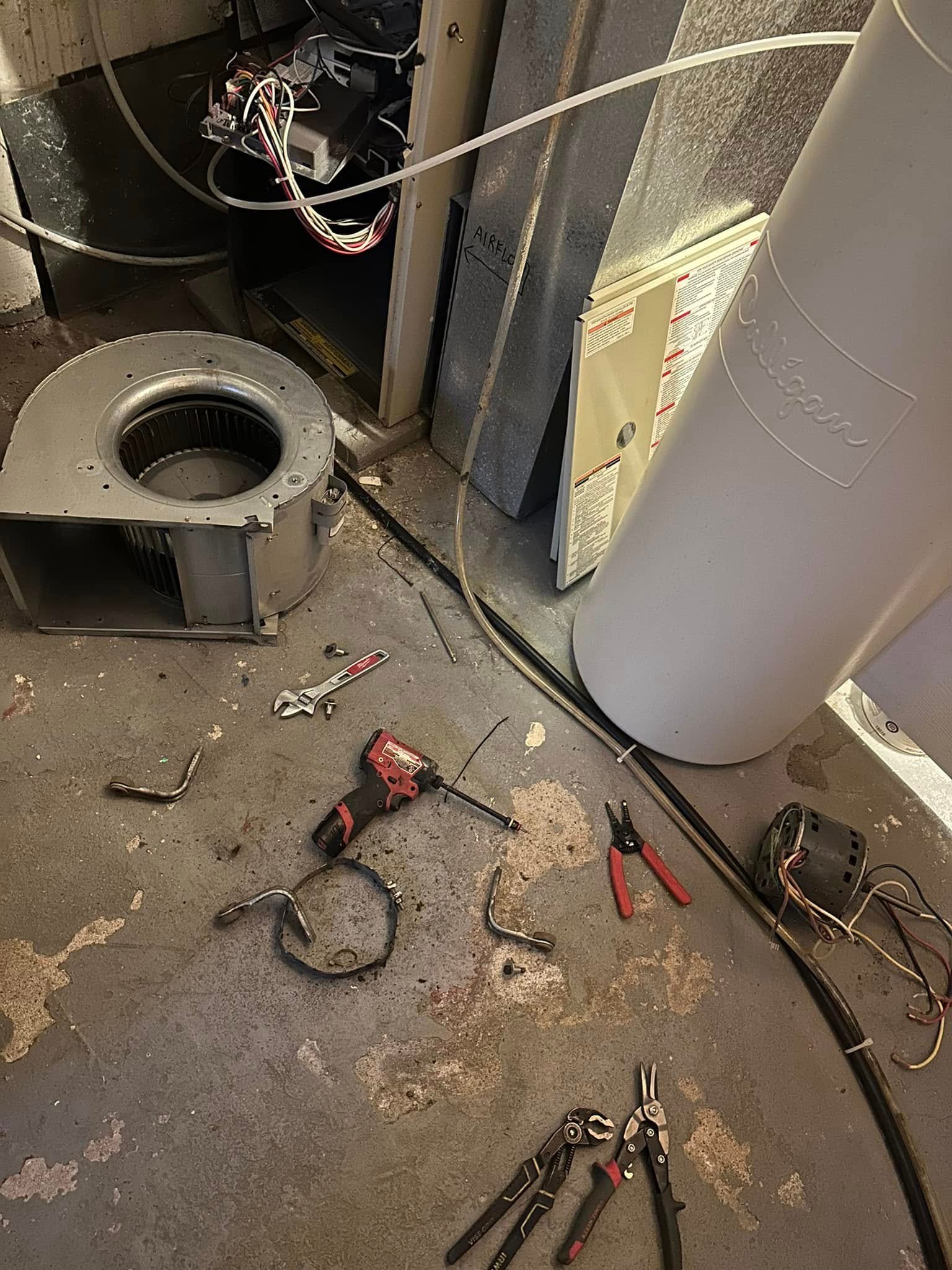 Tools and HVAC parts including blower motor, pliers, and drill on a concrete floor near a water heater.