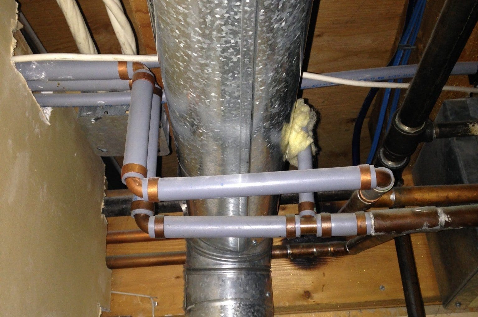 Indoor plumbing with gray pipes, copper fittings, and a central metal duct against a wooden background.