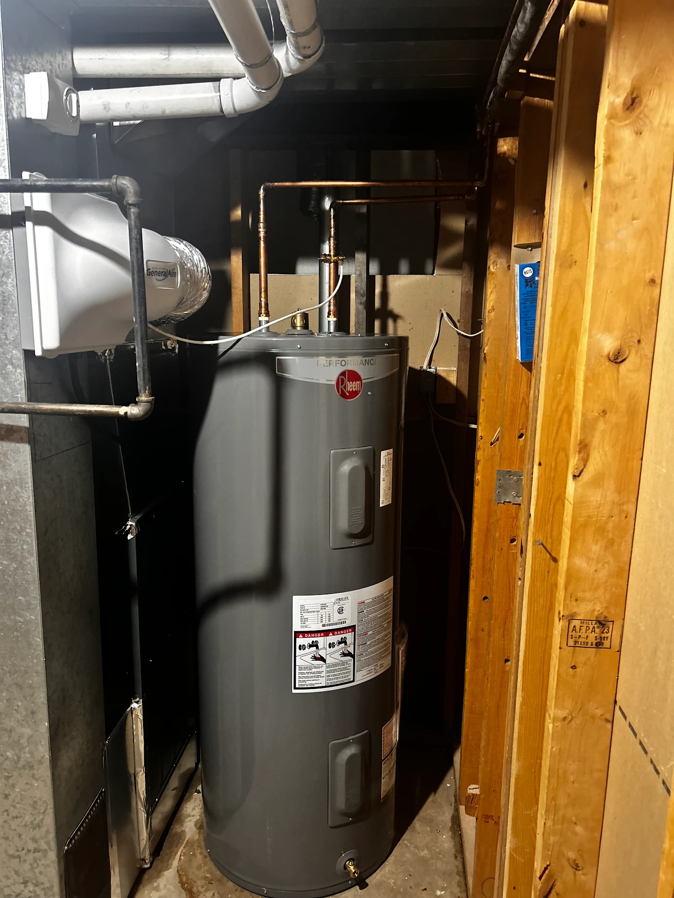 Gray Rheem Performance water heater installed in a basement with copper piping and nearby wooden framing.