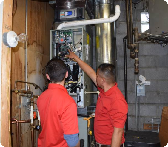 Two plumbers working on a furnace, older plumber showing a younger plumber something in the furnace controls